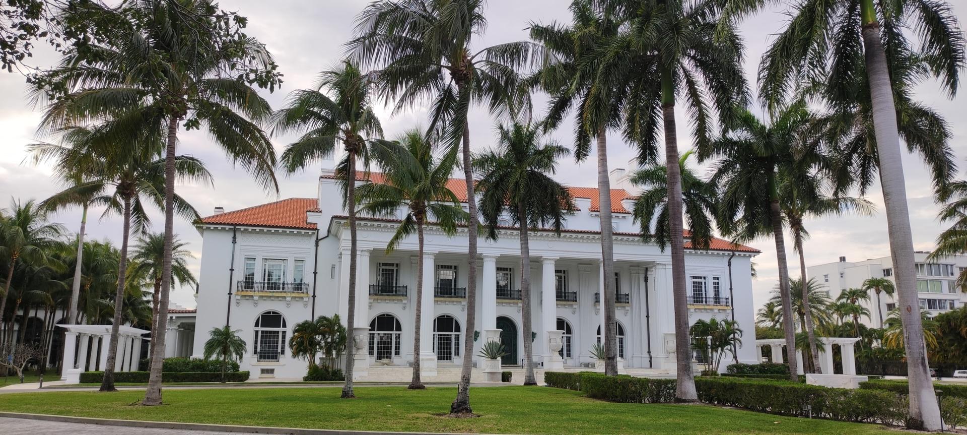Grand white mansion with columns and red roof, framed by tall palm trees.