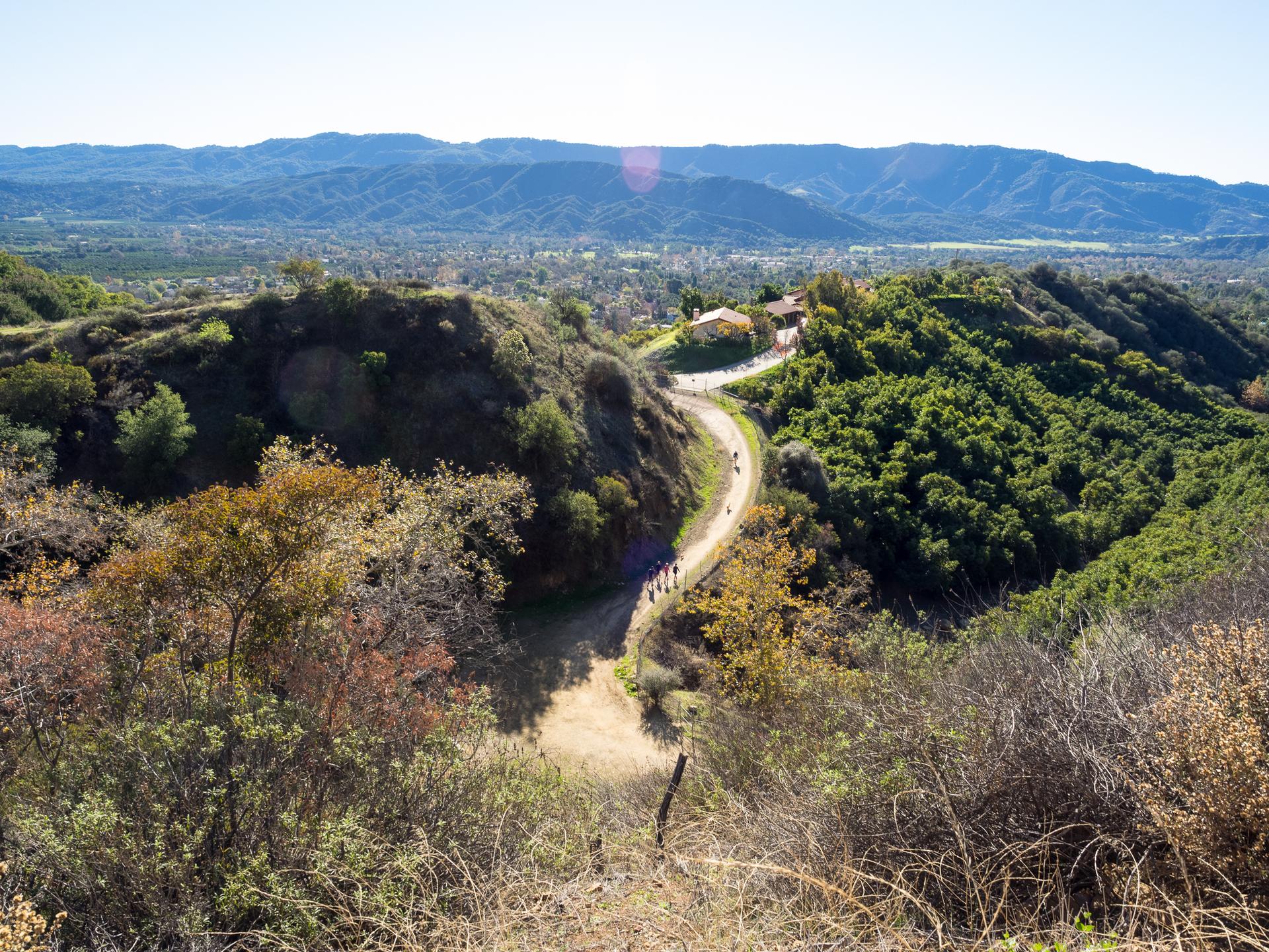A group walks on a winding dirt path through green hills on a sunny day.