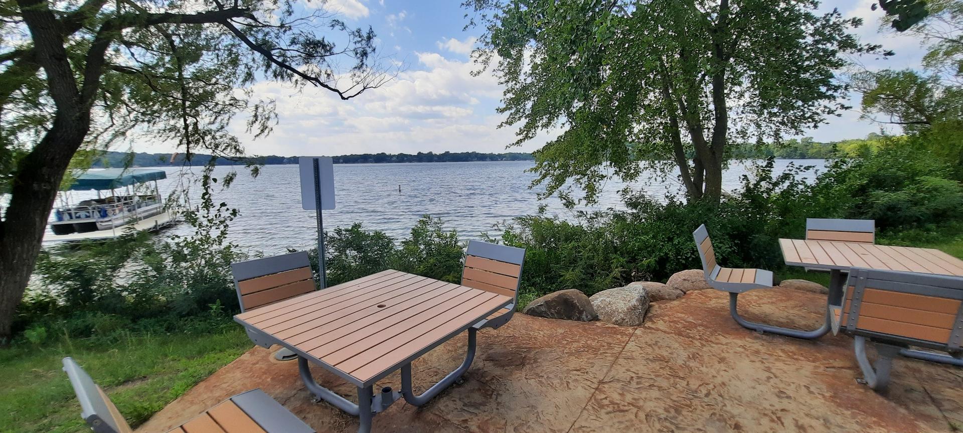 Picnic tables overlook a calm lake with a boat under a partly cloudy sky.