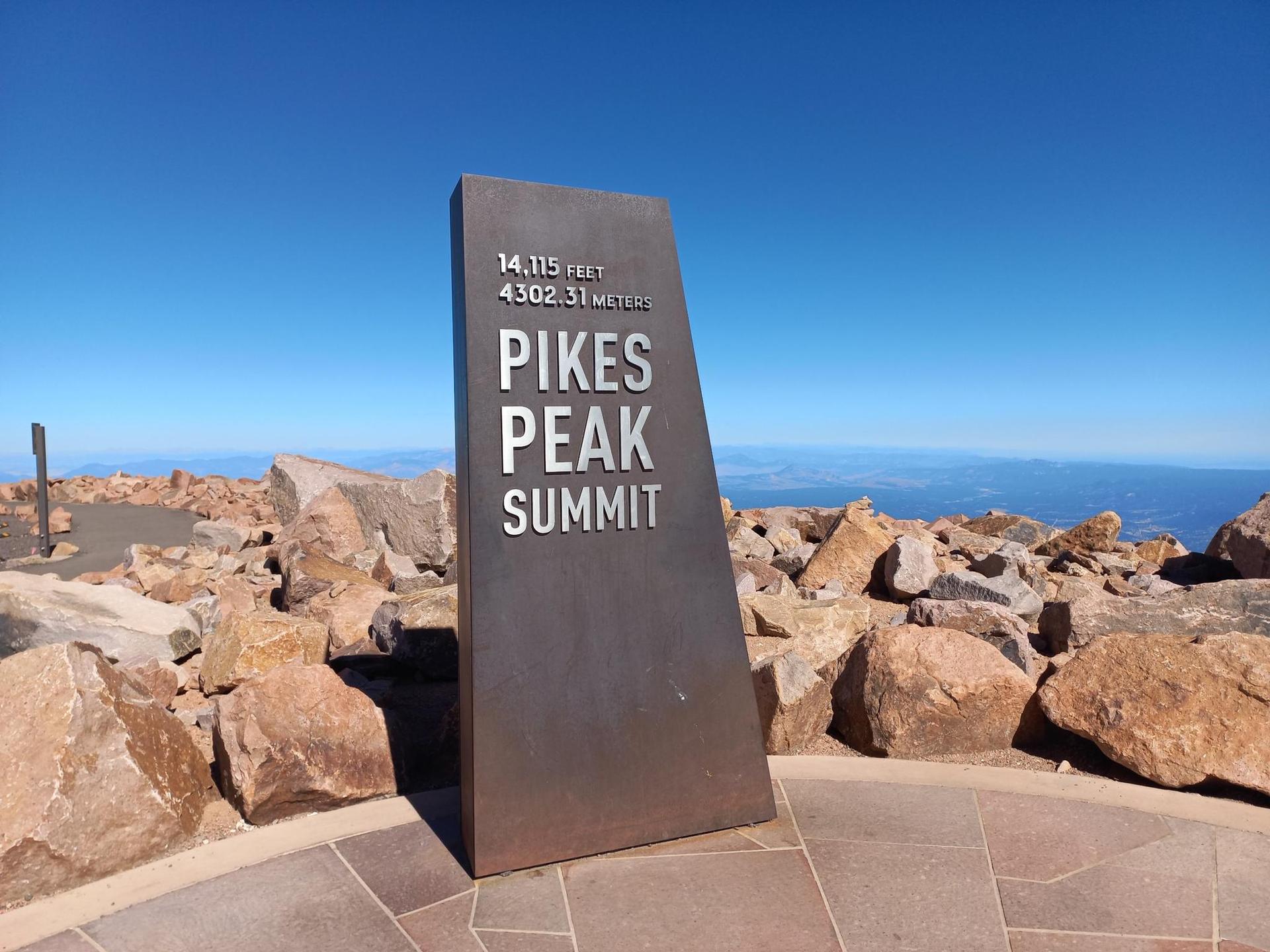 Metal summit marker on a rocky mountain peak under a clear blue sky.
