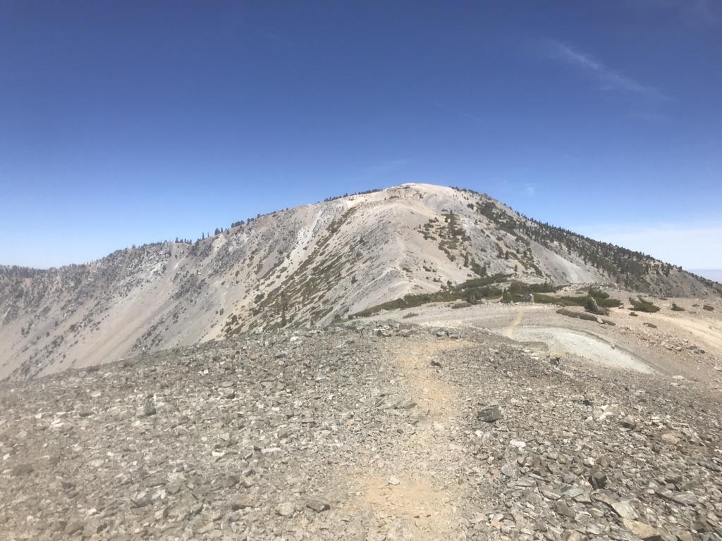 A wide, rocky trail ascends towards a barren mountain peak under clear sky.