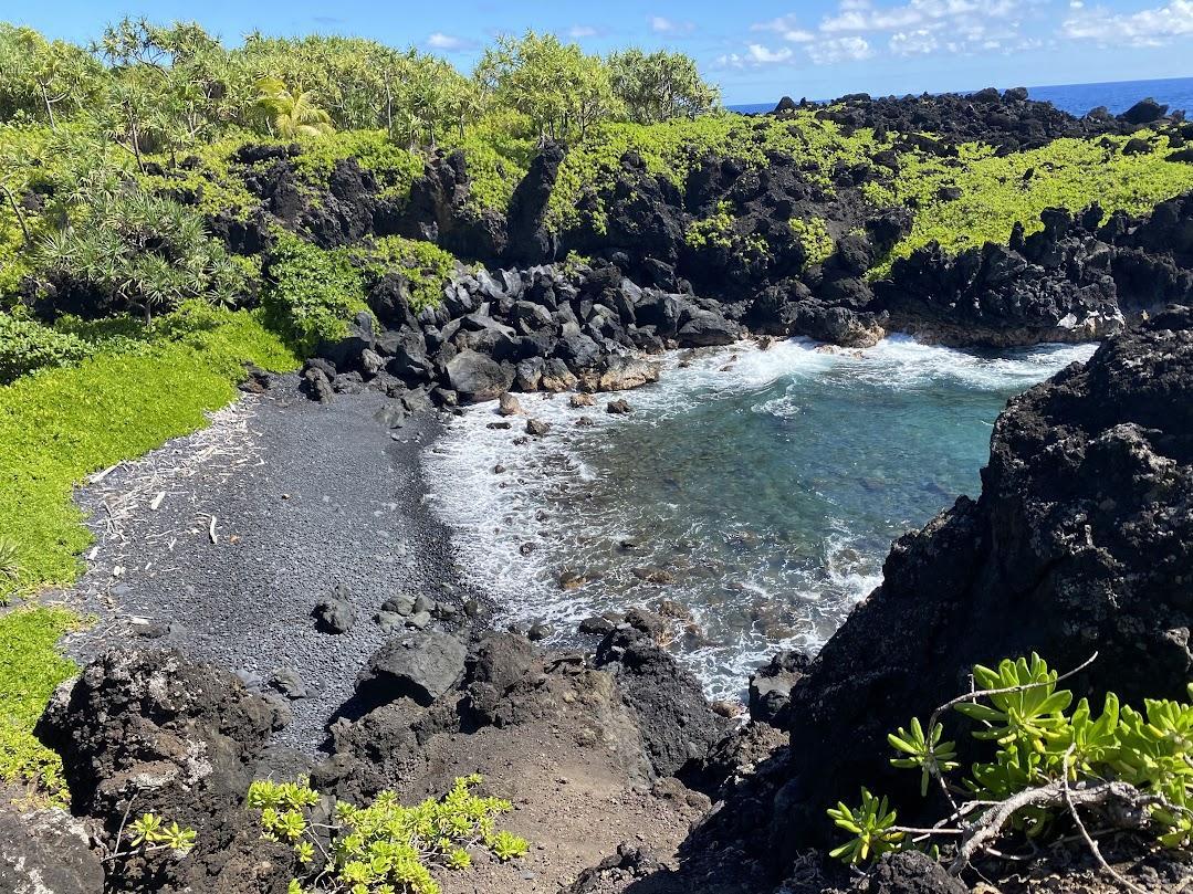 Black sand beach in a rocky cove with lush green vegetation under bright sun.