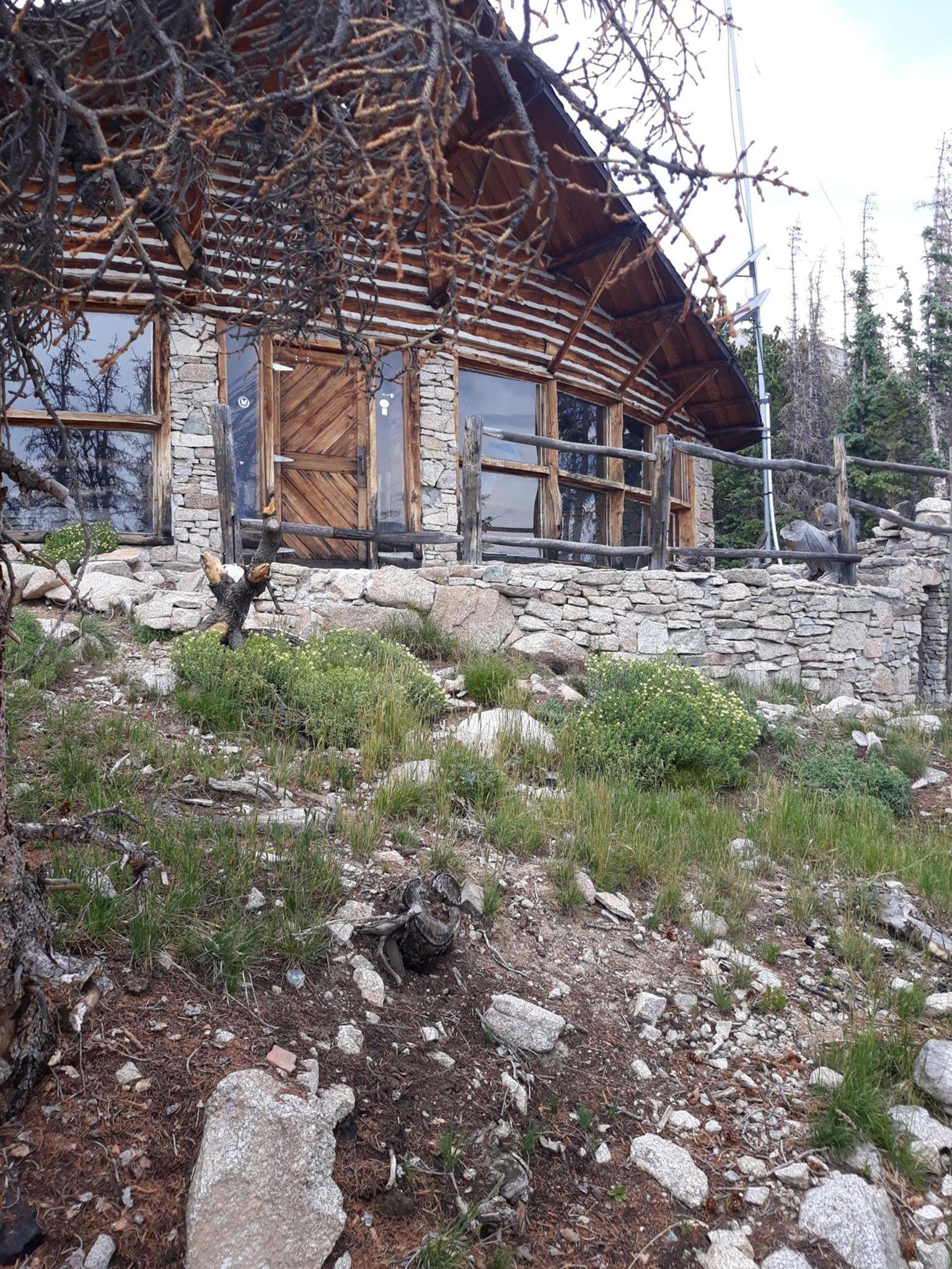 Rustic log and stone cabin on a rocky, grassy hillside under diffused light.