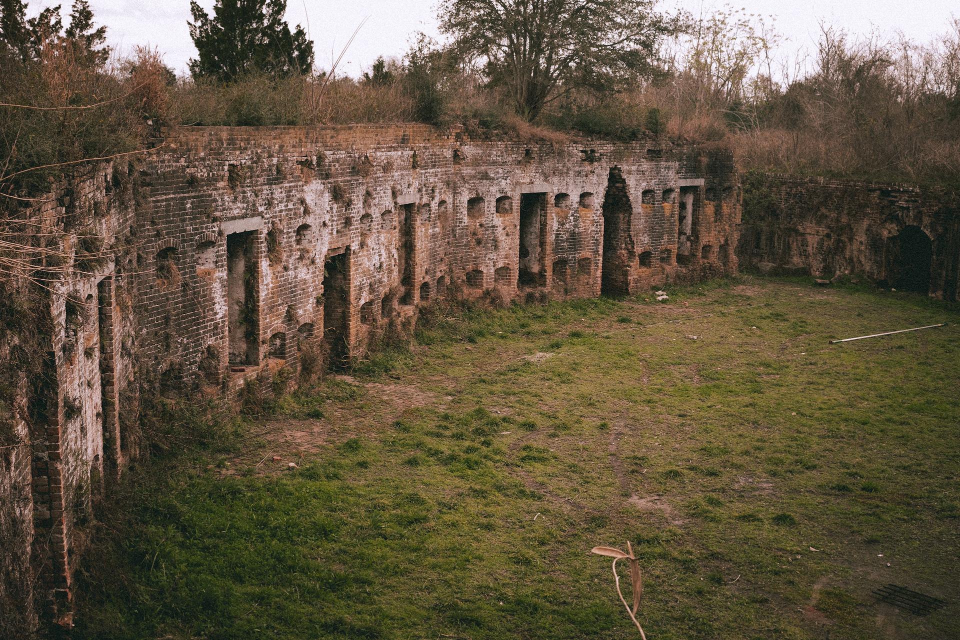 Overgrown ruins of a brick fortification with arched openings and grassy ground.