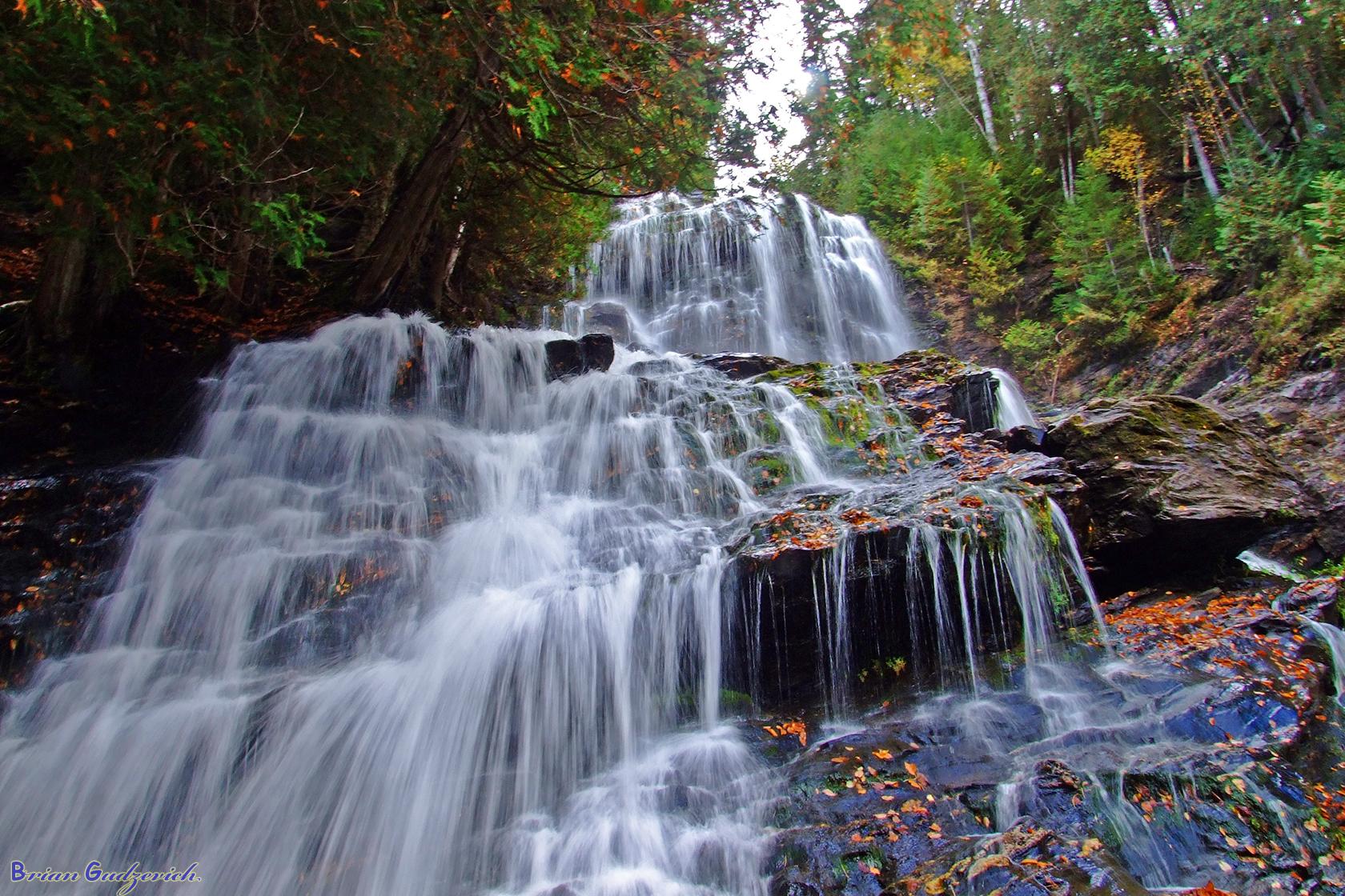 A multi-tiered waterfall cascades over dark rocks amidst autumn trees.