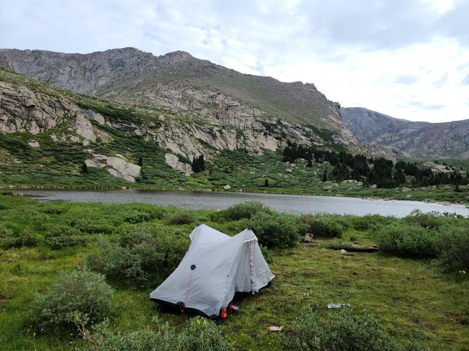 A light-colored tent pitched on a grassy shore next to a mountain lake.