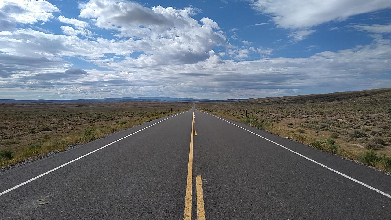 A long, straight paved road stretches through arid scrubland under a cloudy sky.