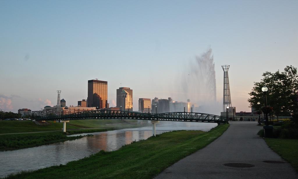 City skyline and bridge over a river with a fountain at sunset.