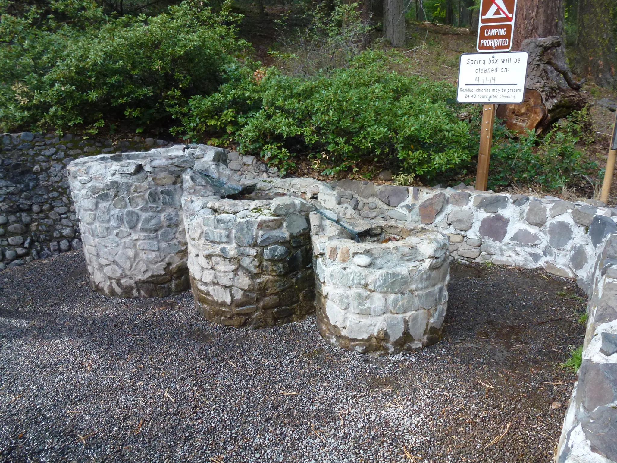 Three stone-lined basins and a sign on gravel, surrounded by green foliage.