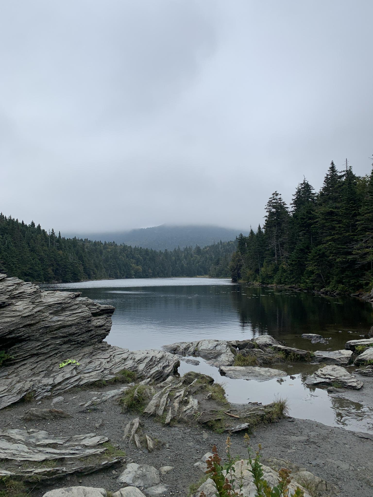 Overcast day at a calm lake surrounded by dense evergreen forests and rocky shores.