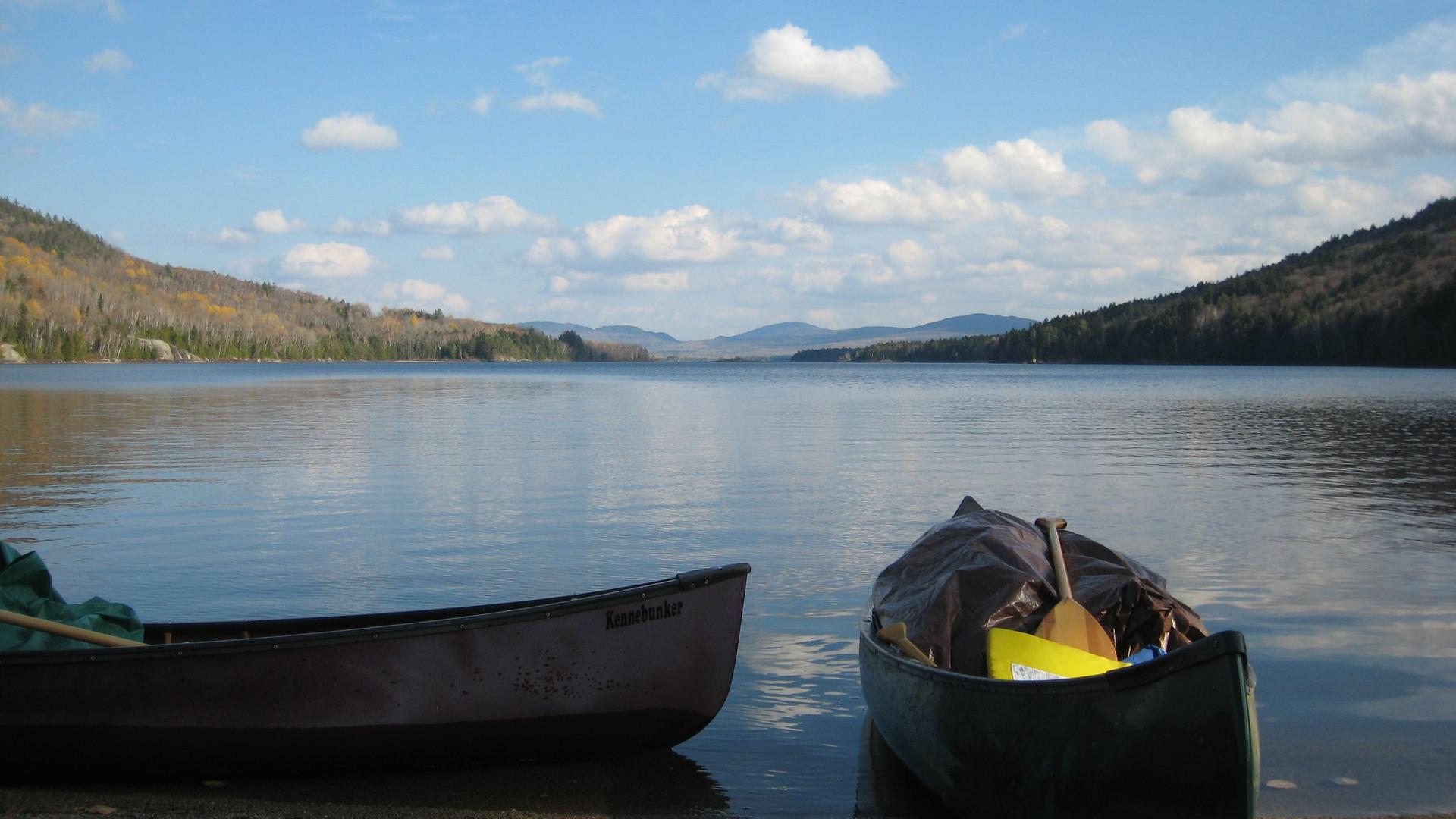 Two canoes on a lake shore with forested mountains under a blue sky.