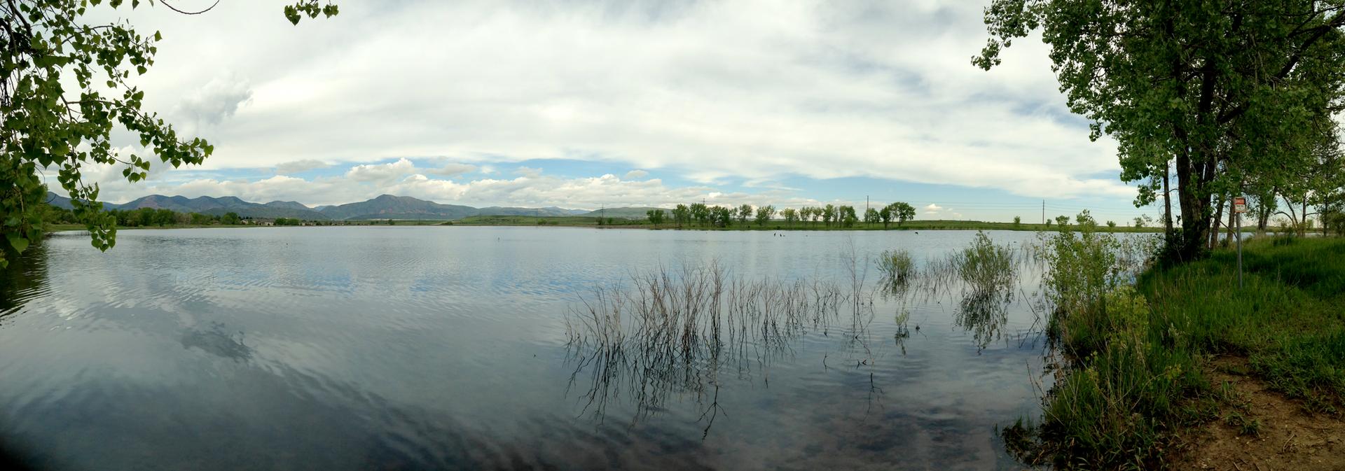 Calm lake reflecting cloudy sky, with distant hills and green trees.