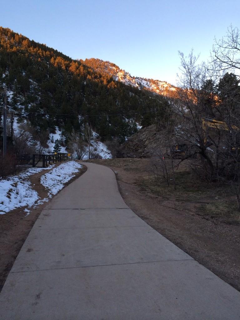 A paved path curves beside a snowy, forested mountain illuminated by golden light.