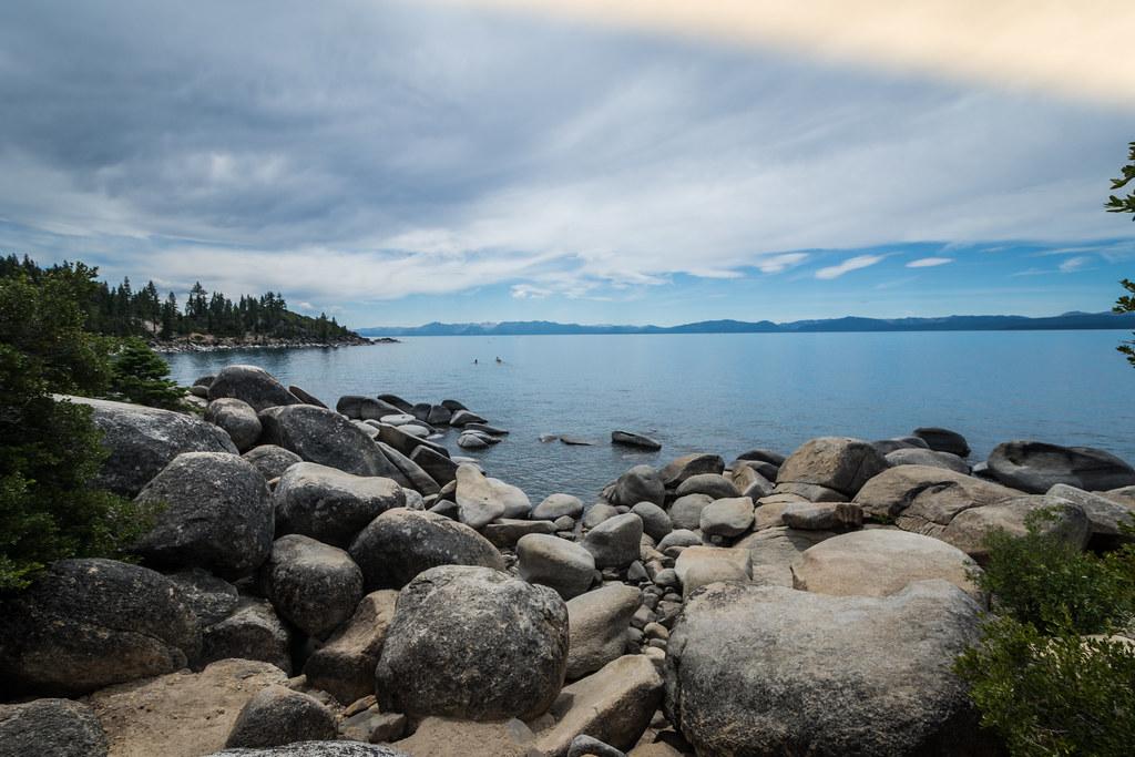 Rocky shoreline with large boulders, vast blue water, and distant mountains under clouds.