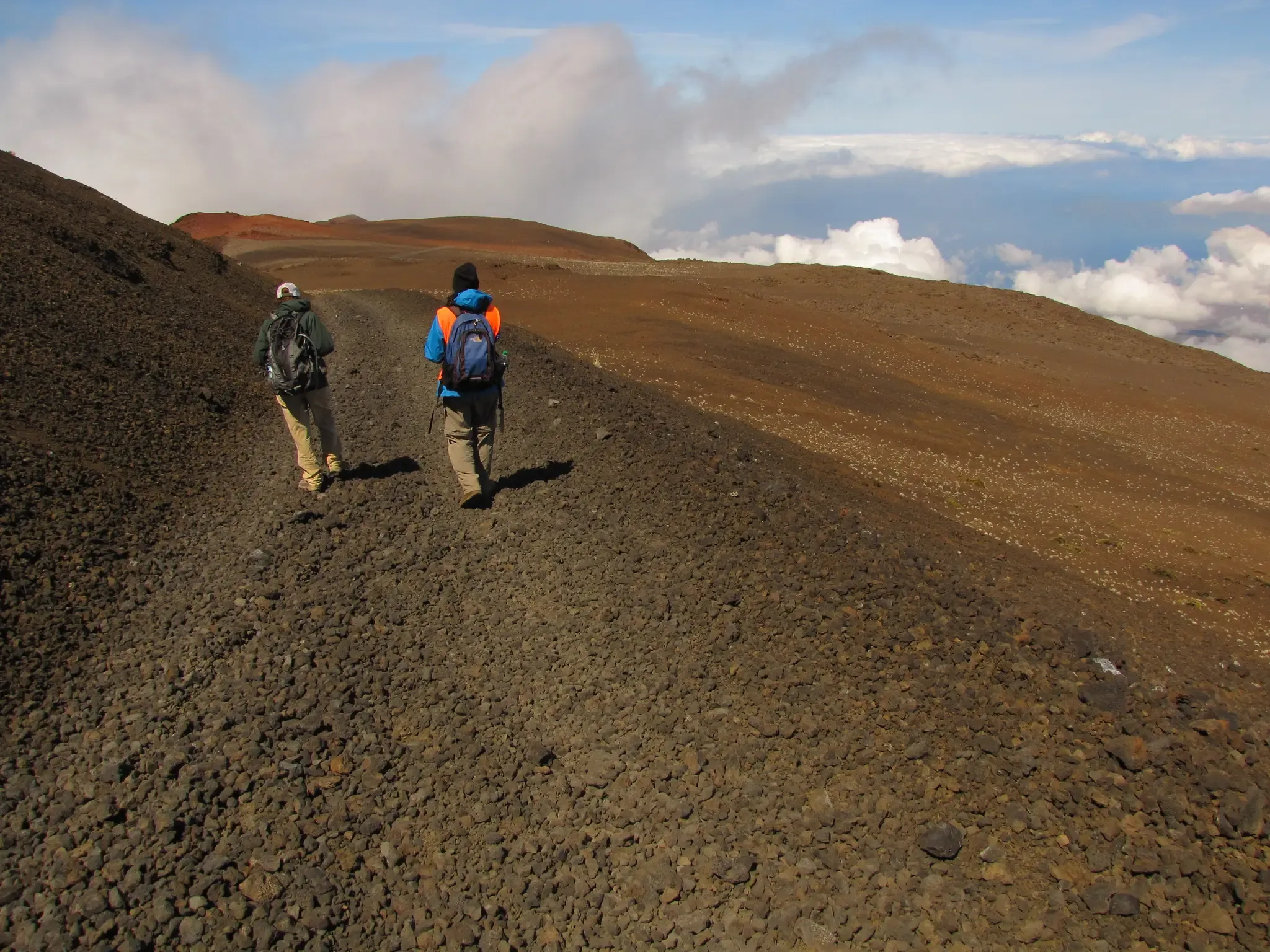 Two hikers walk uphill on a rocky, barren trail under a bright sky. - Maui Skyline Loop