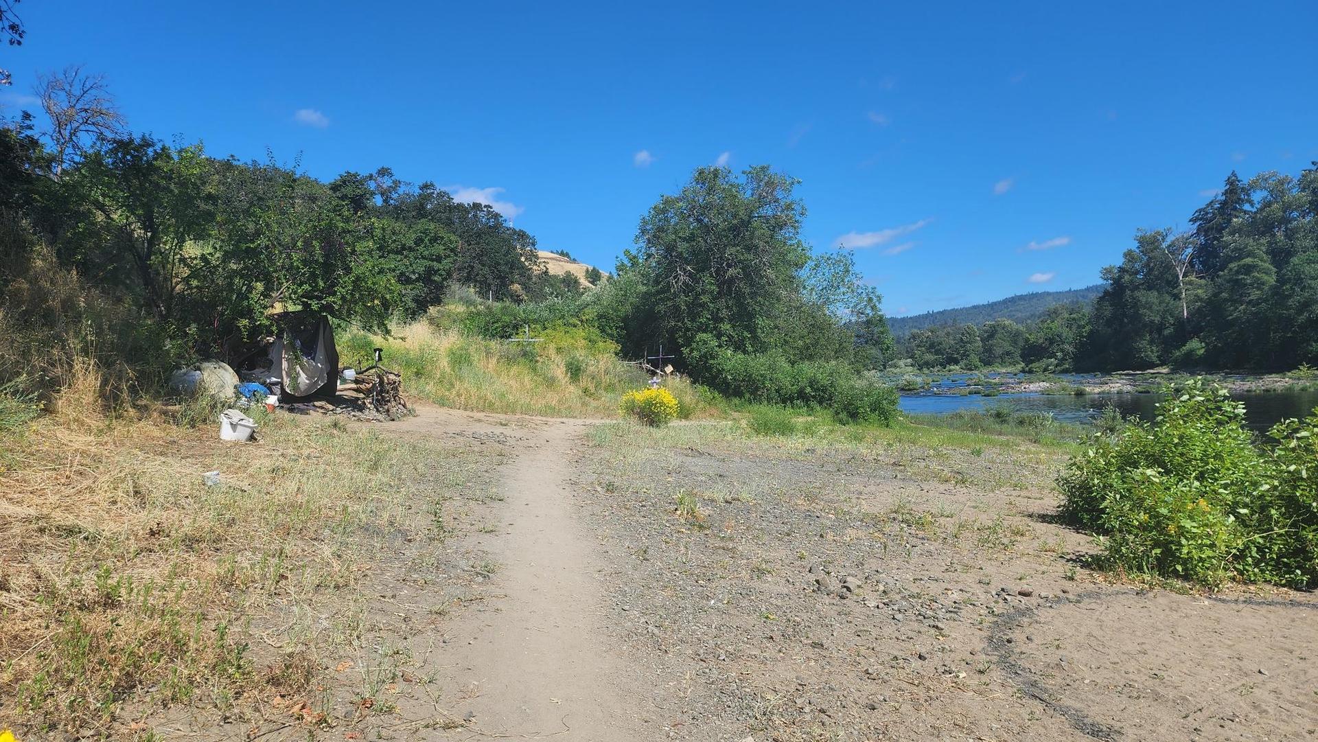 Dirt path leading to a river, with hills and trees under a clear sky.