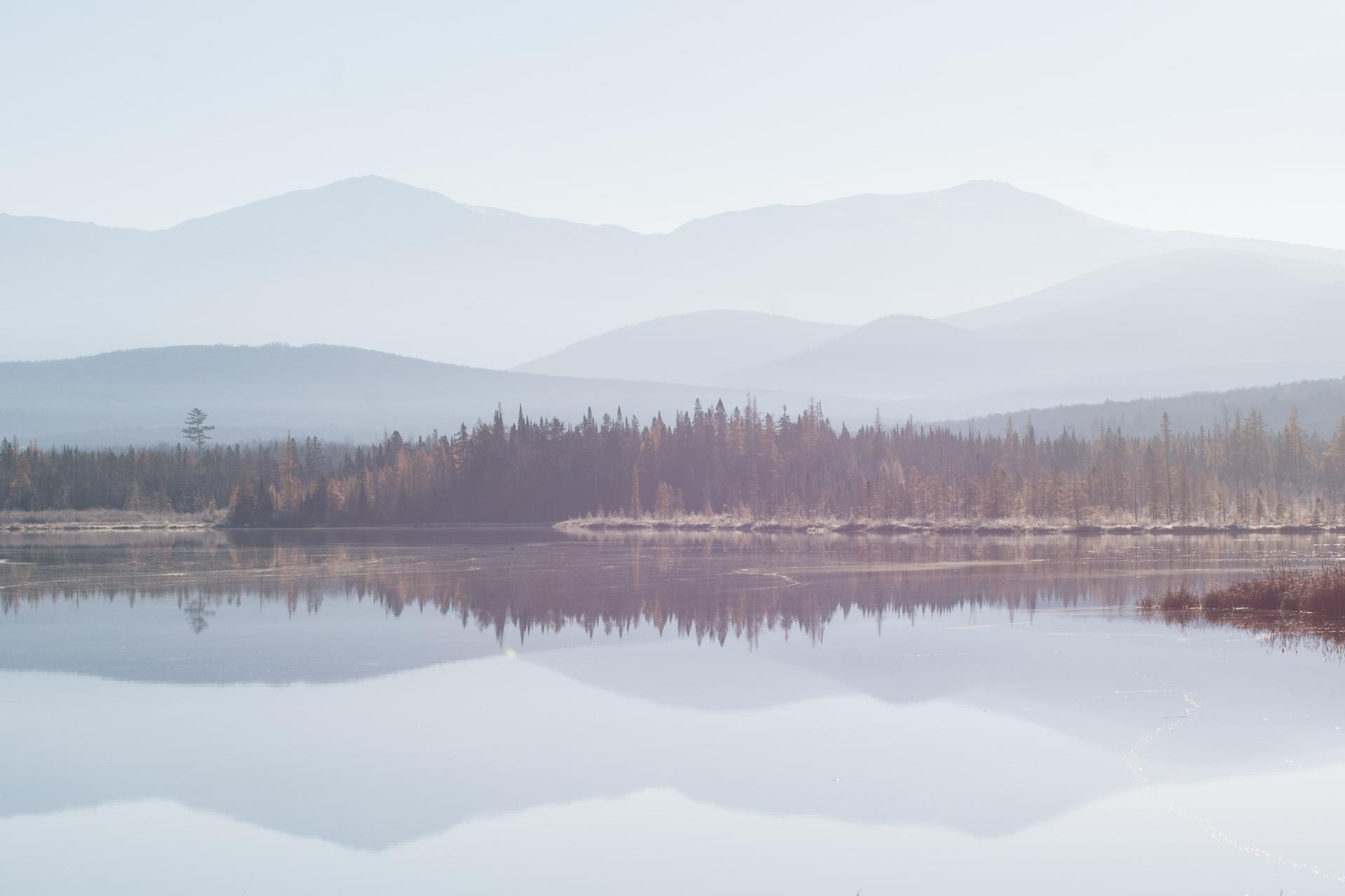 Calm lake reflecting trees and hazy mountains under soft light.