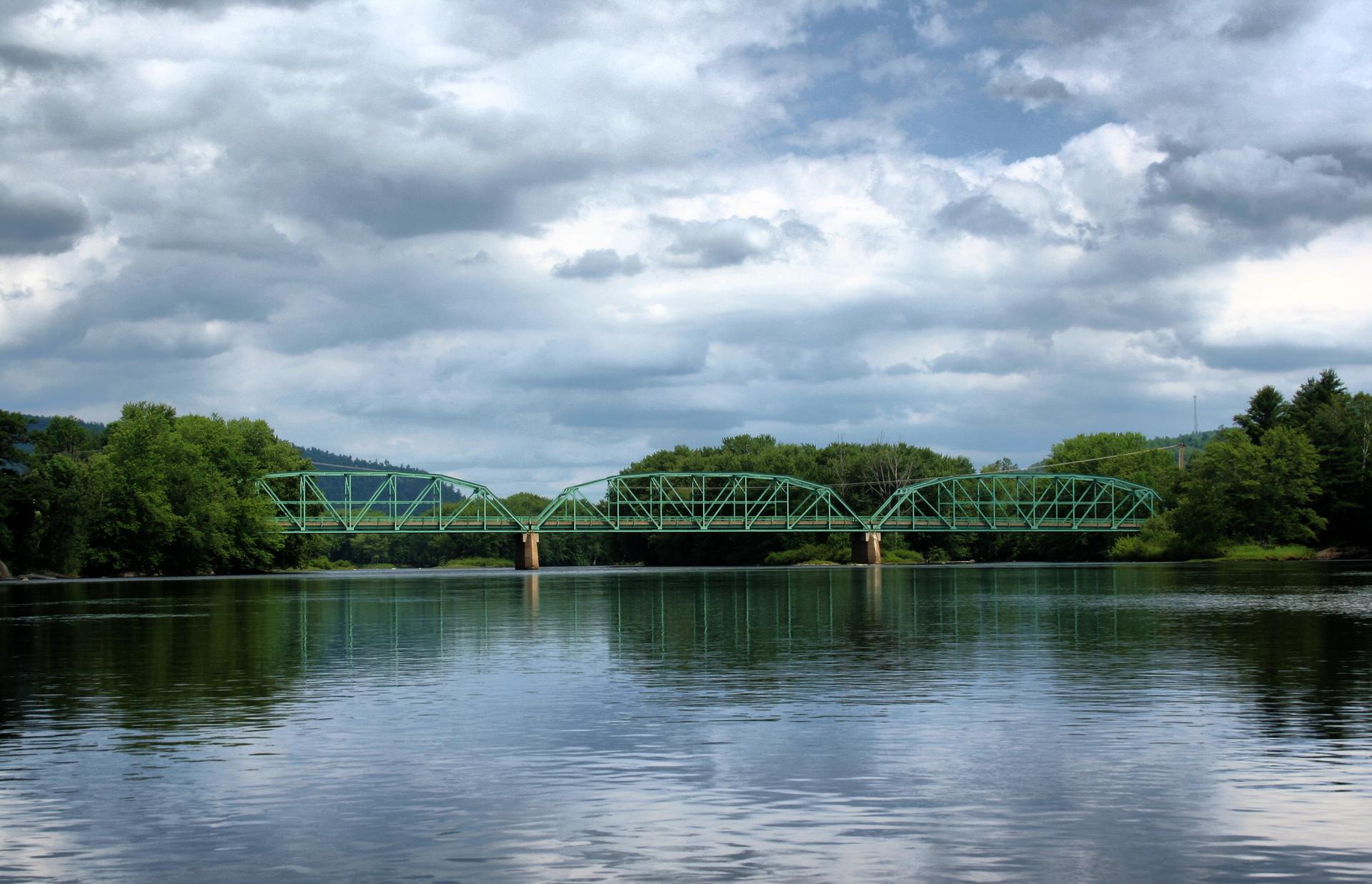 Green truss bridge spans a wide river under a cloudy sky, surrounded by trees.