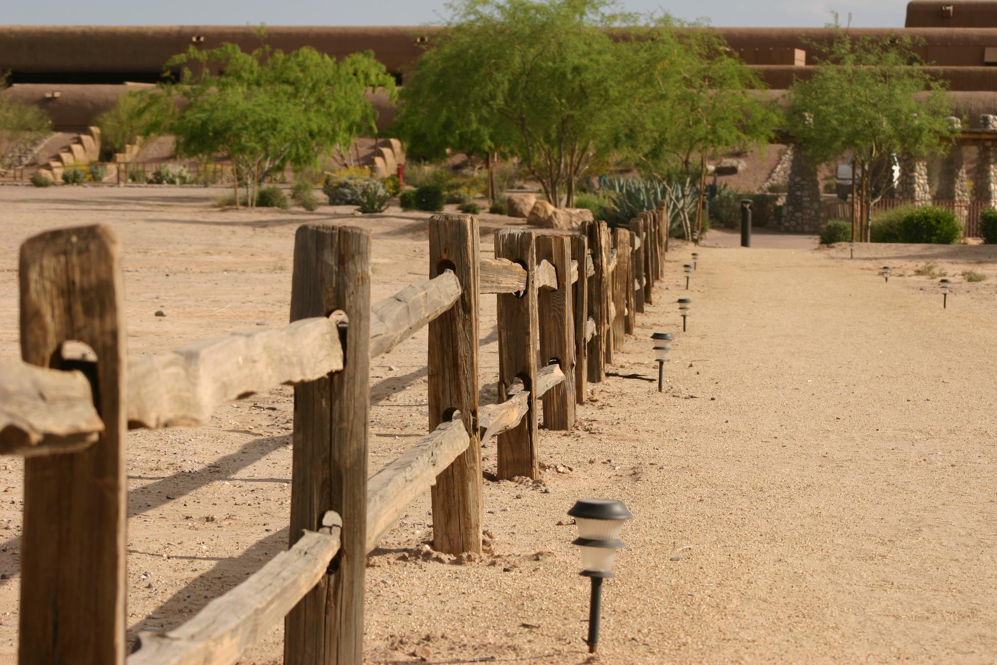 A wooden fence lines a sandy path with solar lights under bright daylight.