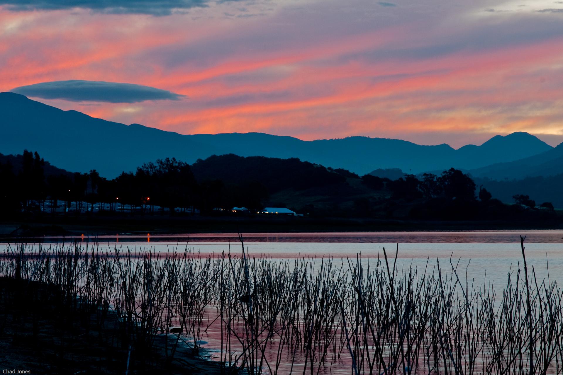 Vibrant pink and orange sunset over a lake with silhouetted mountains and reeds.
