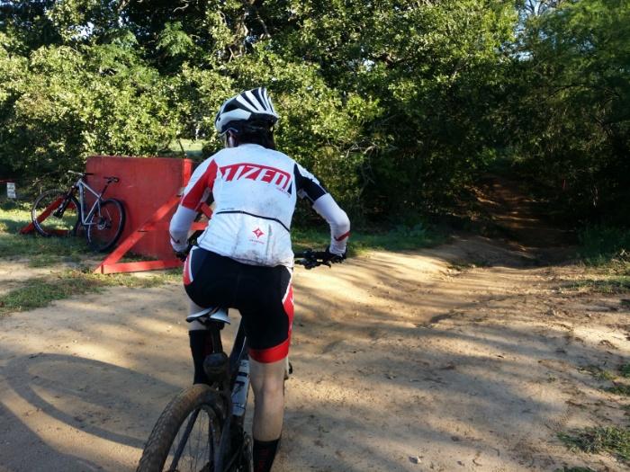 Cyclist on a dirt path, wearing a helmet, surrounded by green trees.