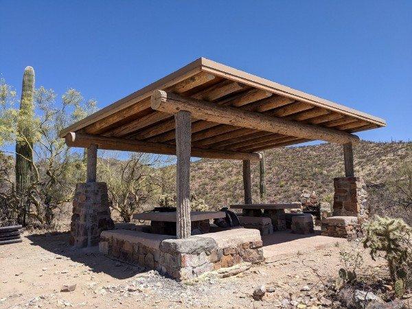 Wooden ramada with stone pillars and benches in a sunny desert setting.