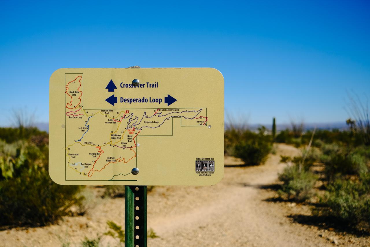 A trail map sign stands on a dirt path under a bright blue sky.
