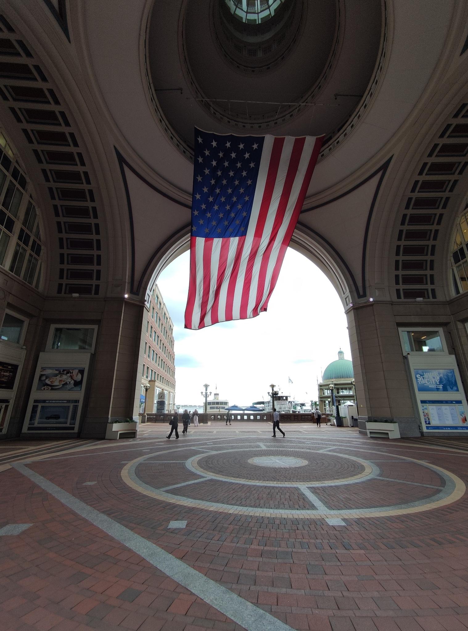 Large American flag hanging in an arched hall with people walking below.