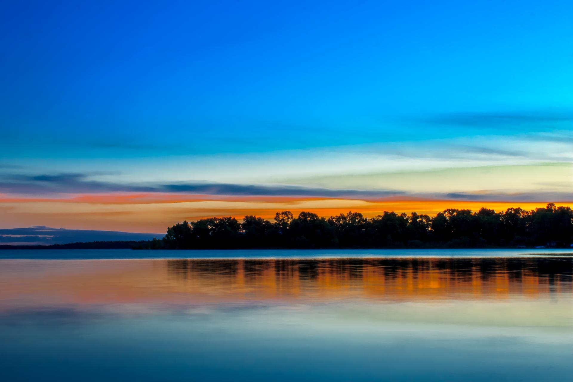 Vibrant sunset over calm water, reflecting colorful sky and distant trees.