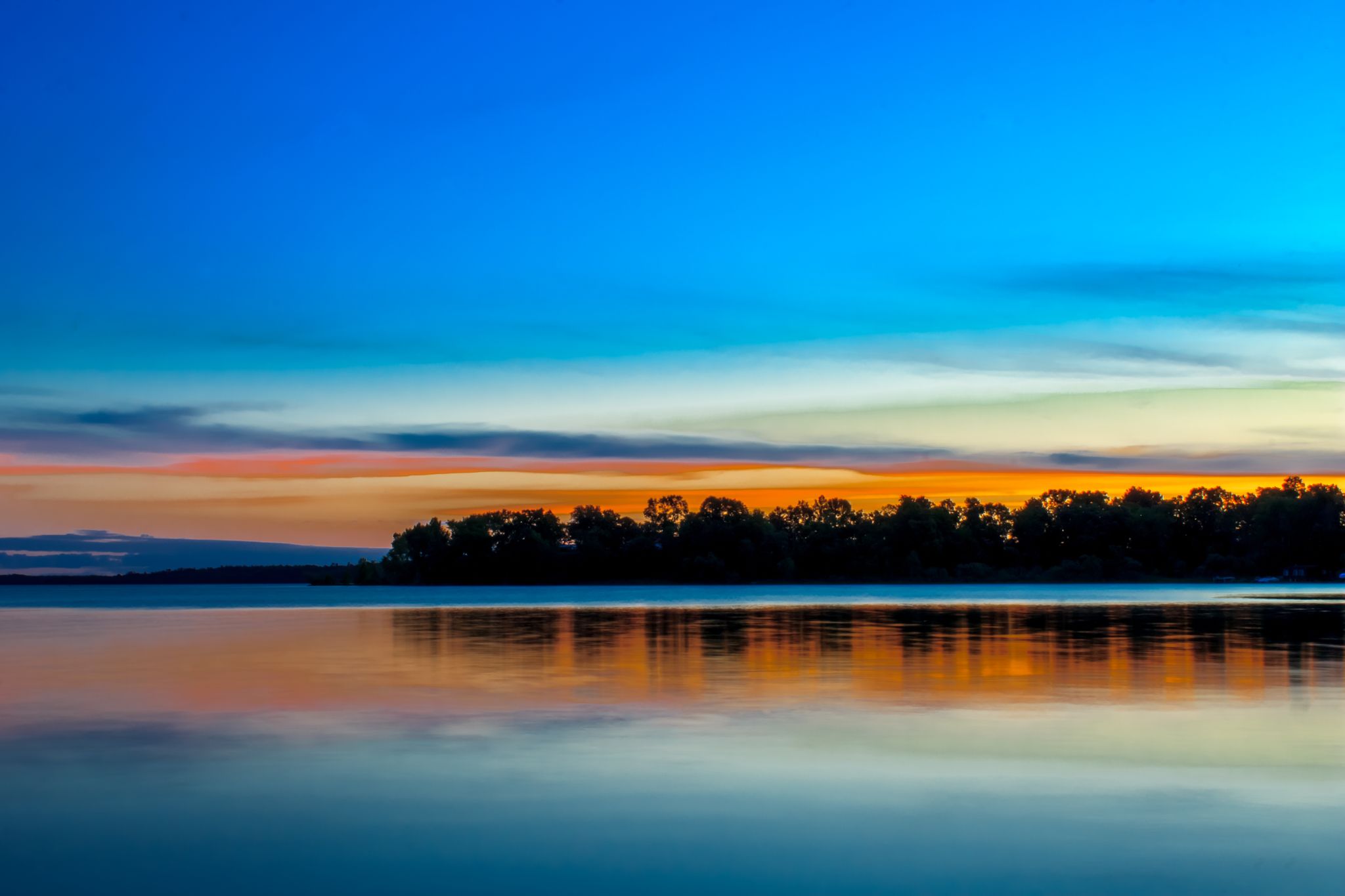 Vibrant sunset over calm water, reflecting colorful sky and distant trees. - Migizi Trail