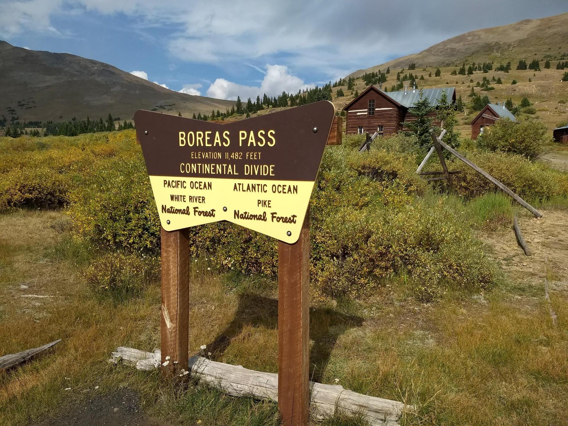 Brown wooden sign on a grassy hillside with distant buildings under bright sky.