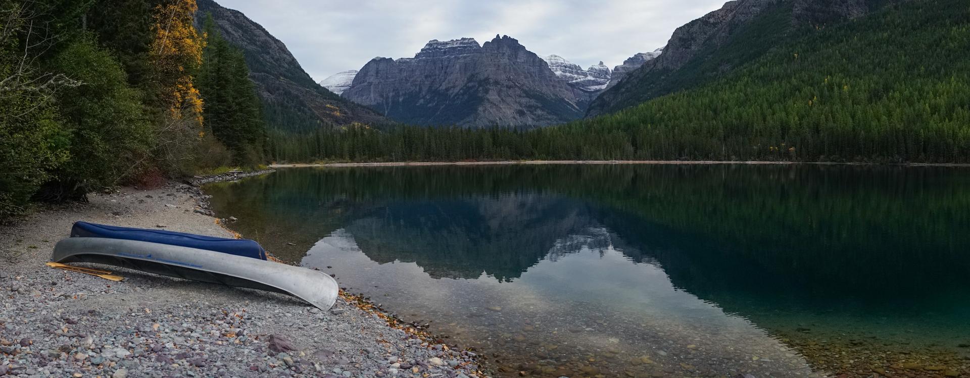 Two canoes on a rocky shore by a calm lake reflecting mountains.