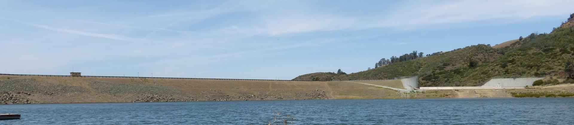 A wide dam and reservoir under a bright blue sky with distant hills.