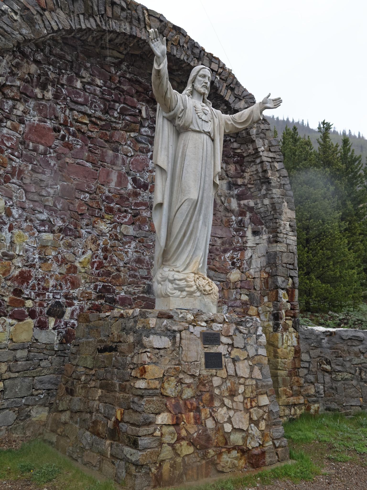White religious statue with outstretched arms against a stone archway and trees.