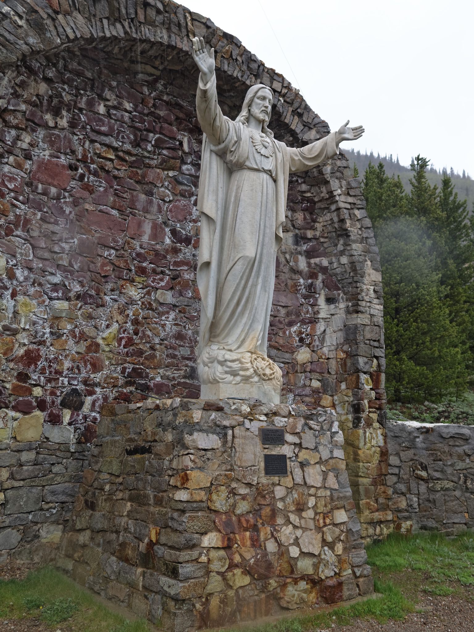 White religious statue with outstretched arms against a stone archway and trees. - Rainbow Trail