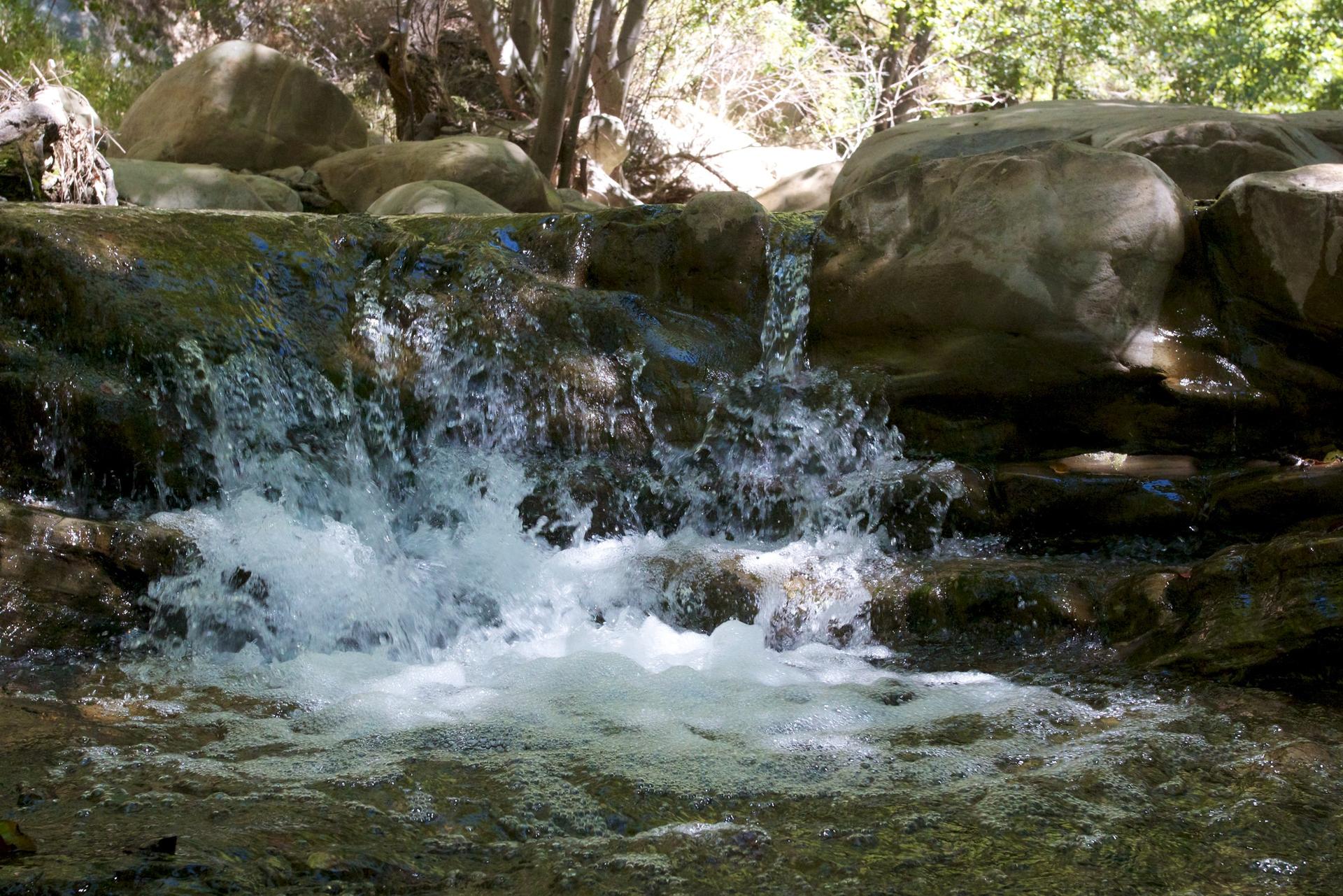 Cascading water over rocks in a shaded stream with lush green surroundings.