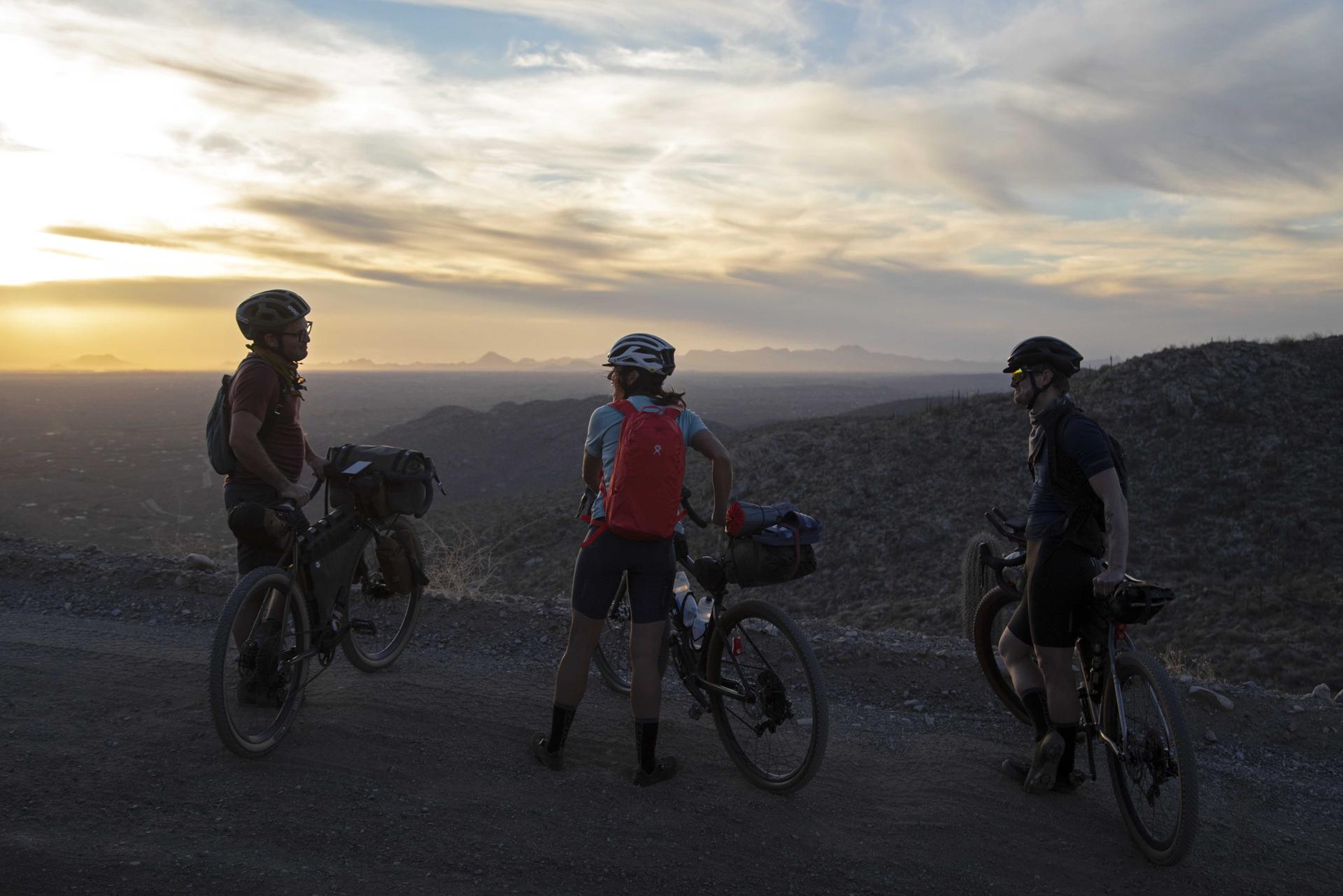 Three cyclists on a dirt road watching a beautiful sunset over hills.