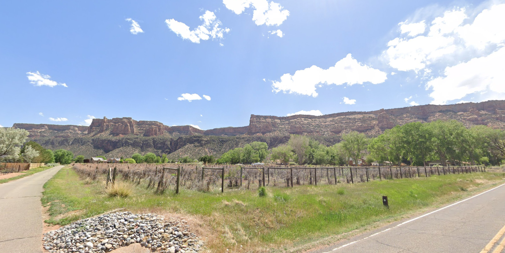 Rural road beside a fenced field, with large flat-topped mesas in the background.