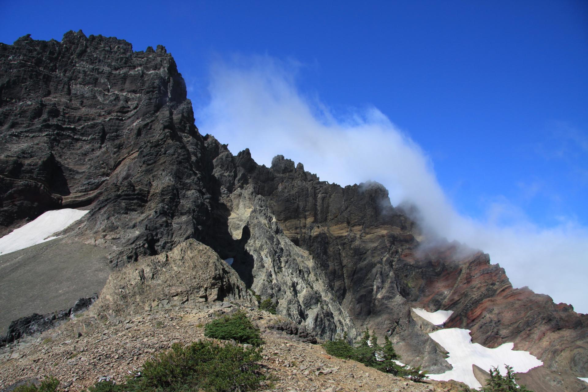 Rugged rocky mountain face with snow and clouds under a bright blue sky.