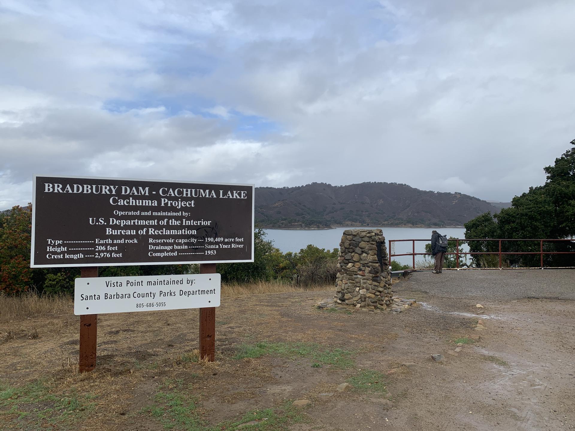 Informational sign overlooking a lake with hills and a person under cloudy sky.