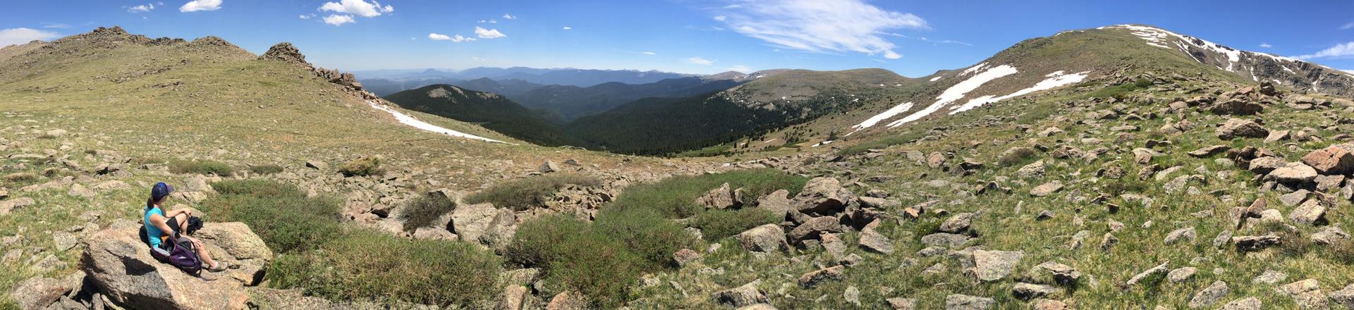 Person sits on rocks overlooking a vast, sunny mountain valley.