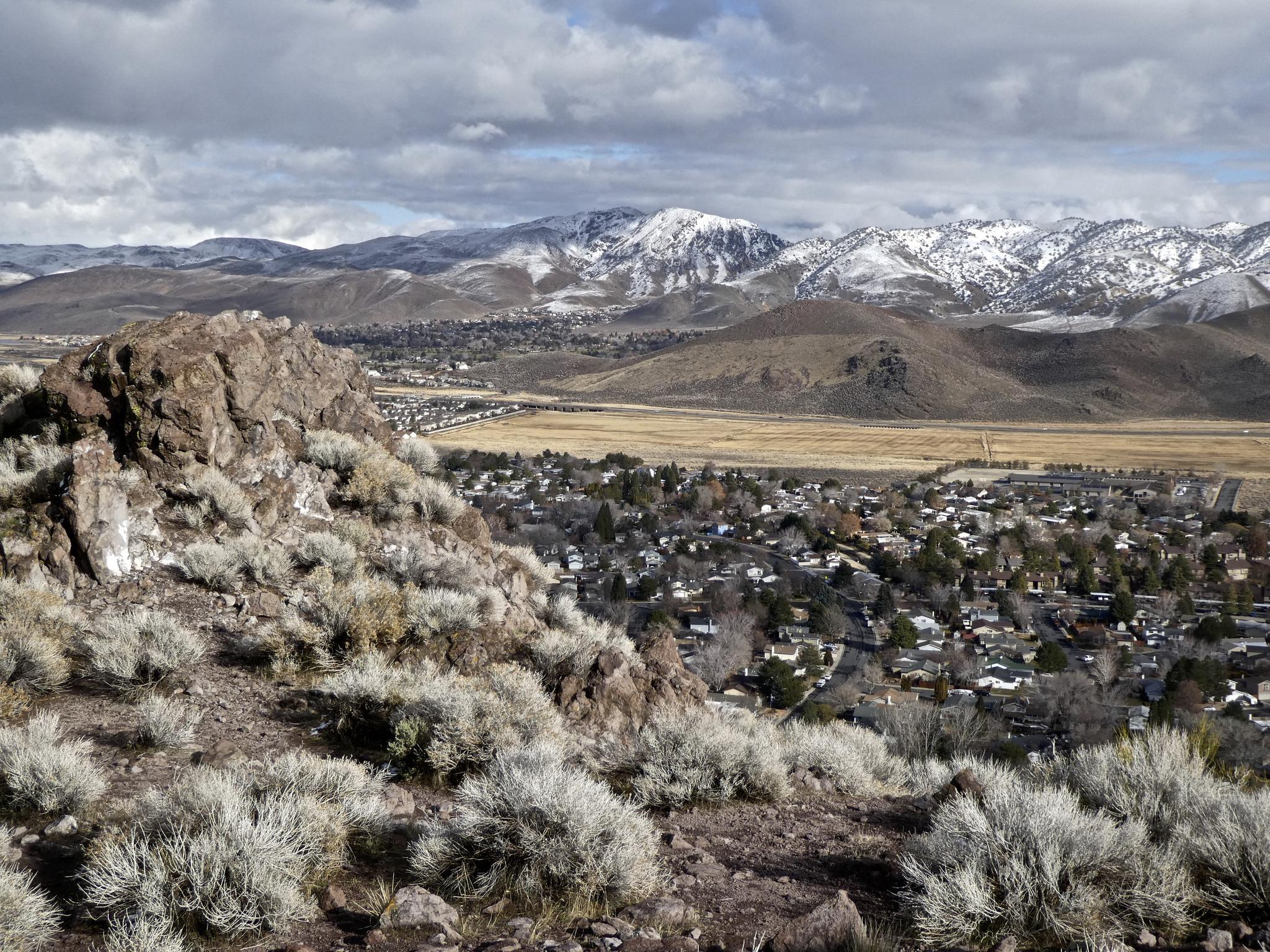 Overcast view from a rocky hill overlooking a town and snow-capped mountains.