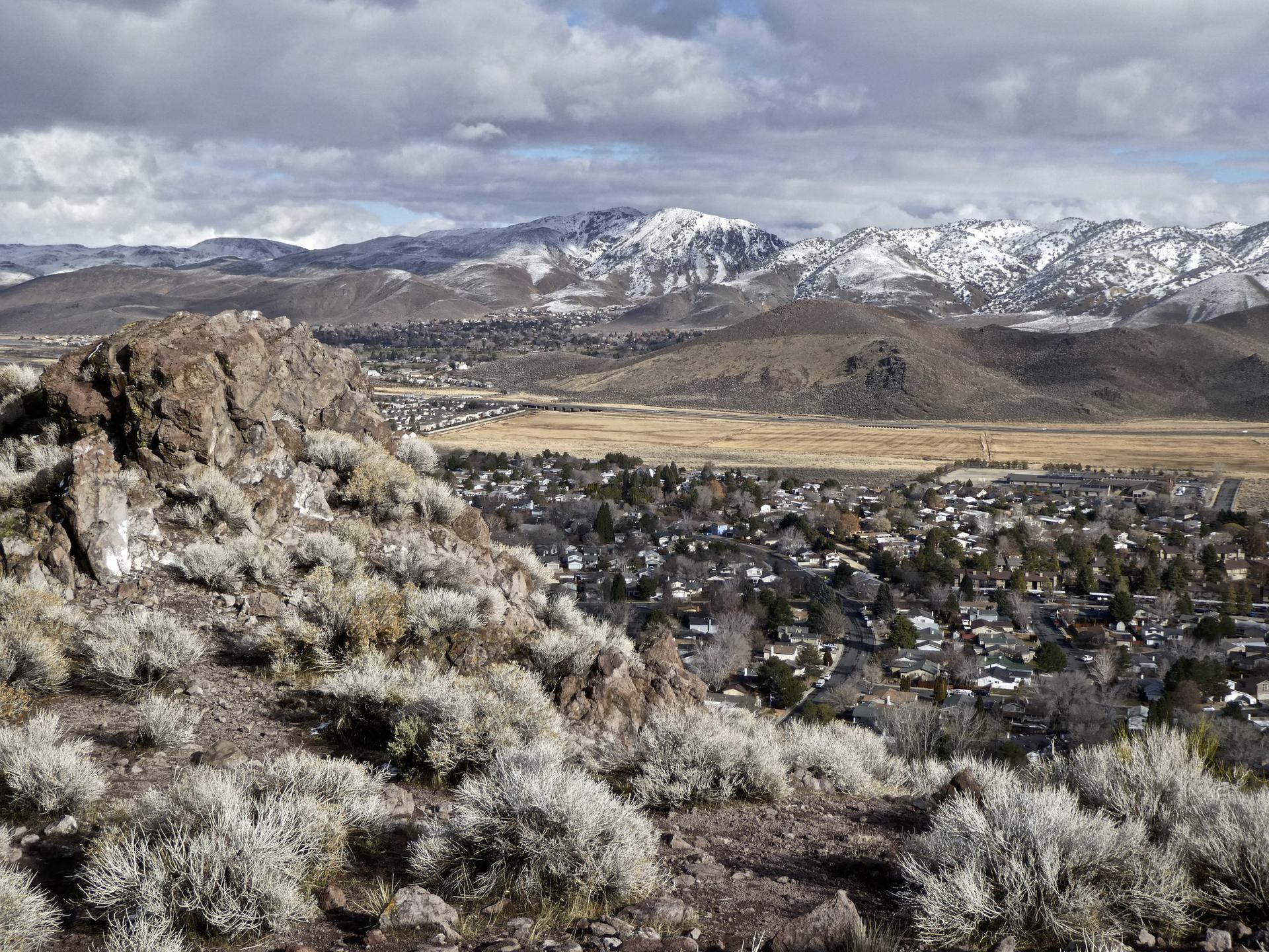 Overcast view from a rocky hill overlooking a town and snow-capped mountains.
