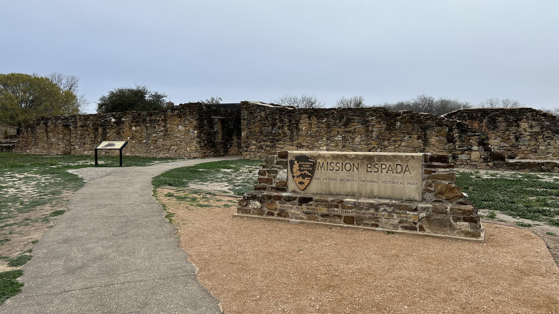 Stone ruins and a sign next to a paved path on an overcast day.