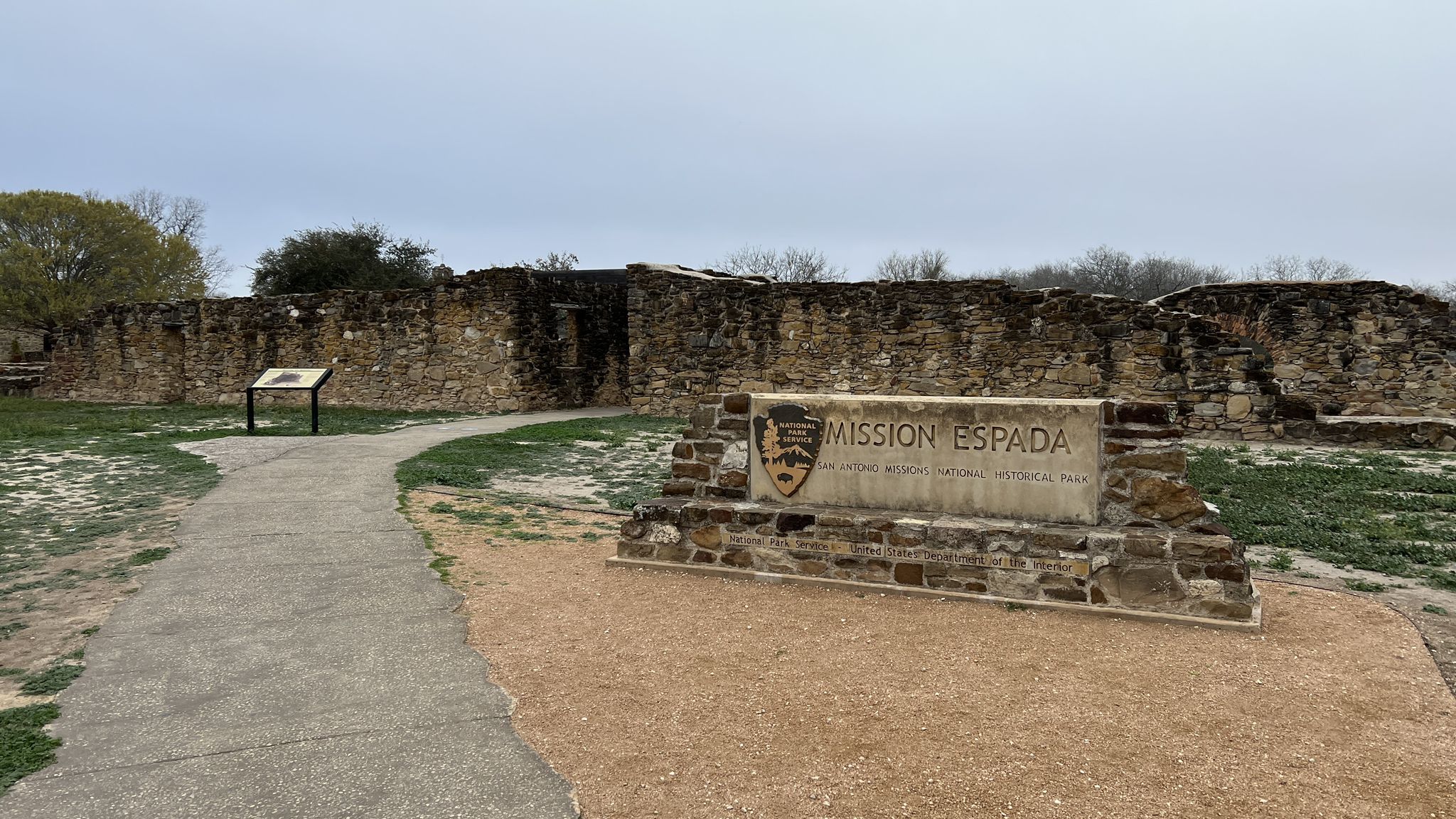 Stone ruins and a sign next to a paved path on an overcast day. - Missions San Juan River Walk