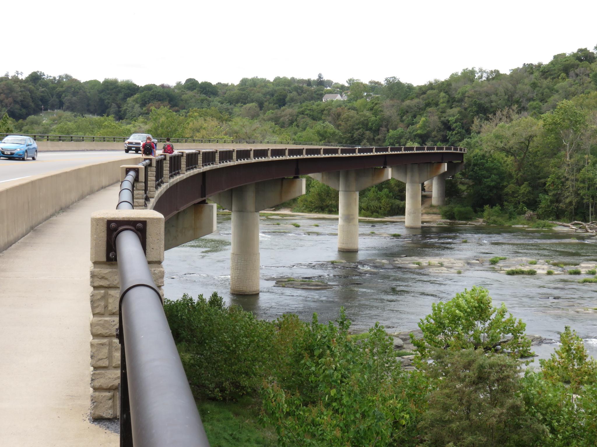 Curved concrete bridge with cars crossing over a rocky river, surrounded by green trees.