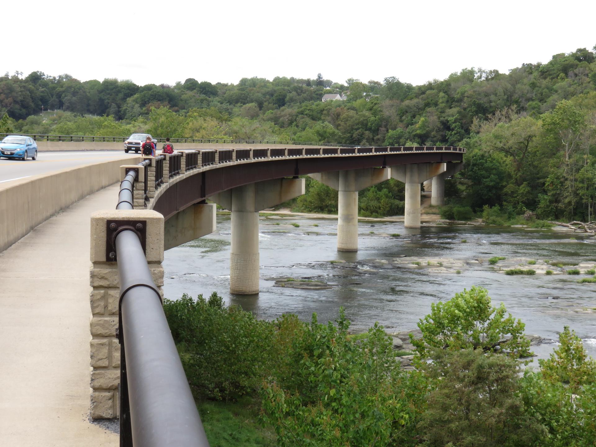 Curved concrete bridge with cars crossing over a rocky river, surrounded by green trees.