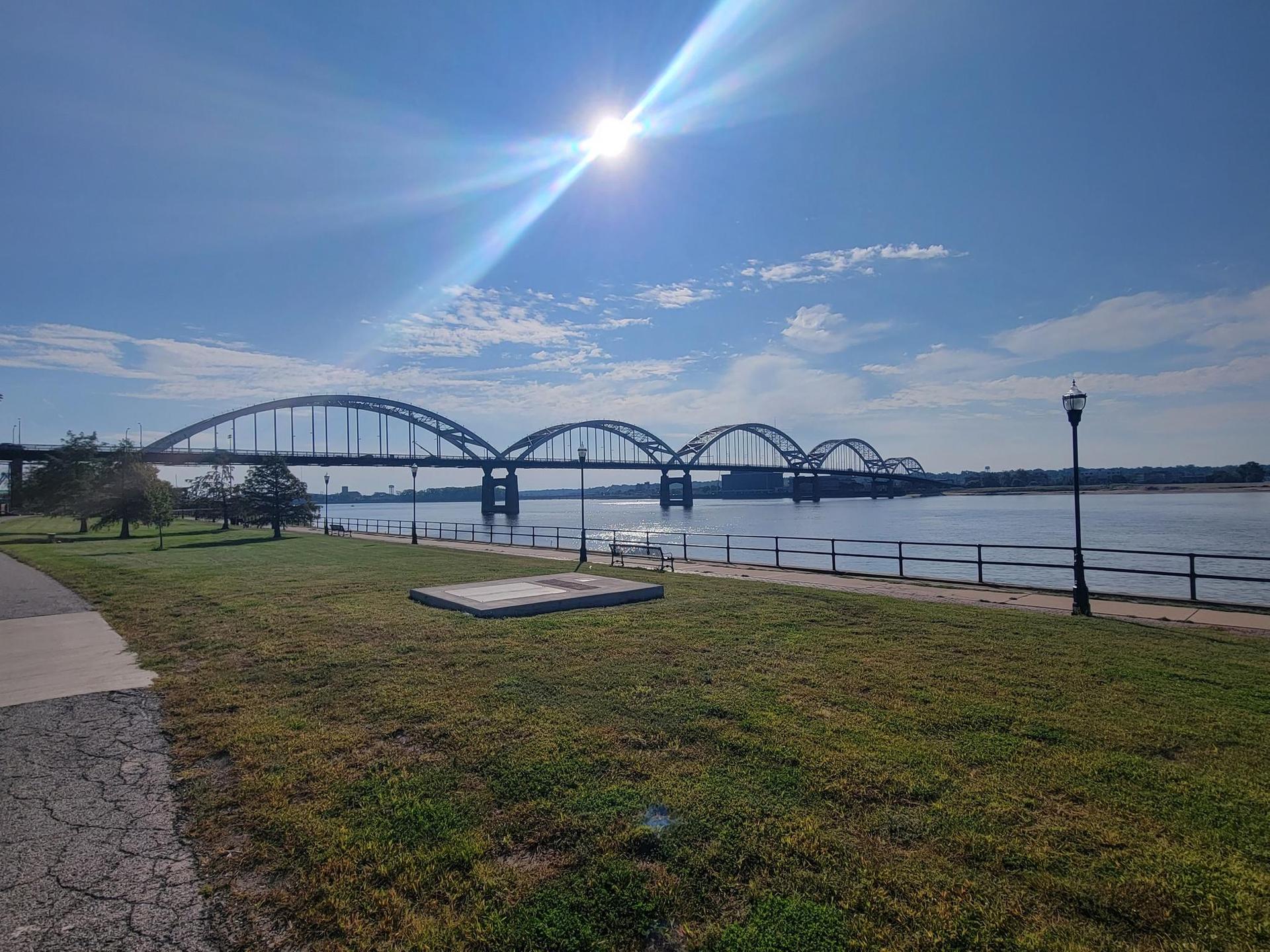 A large arched bridge spans a wide river under a bright sunny sky.