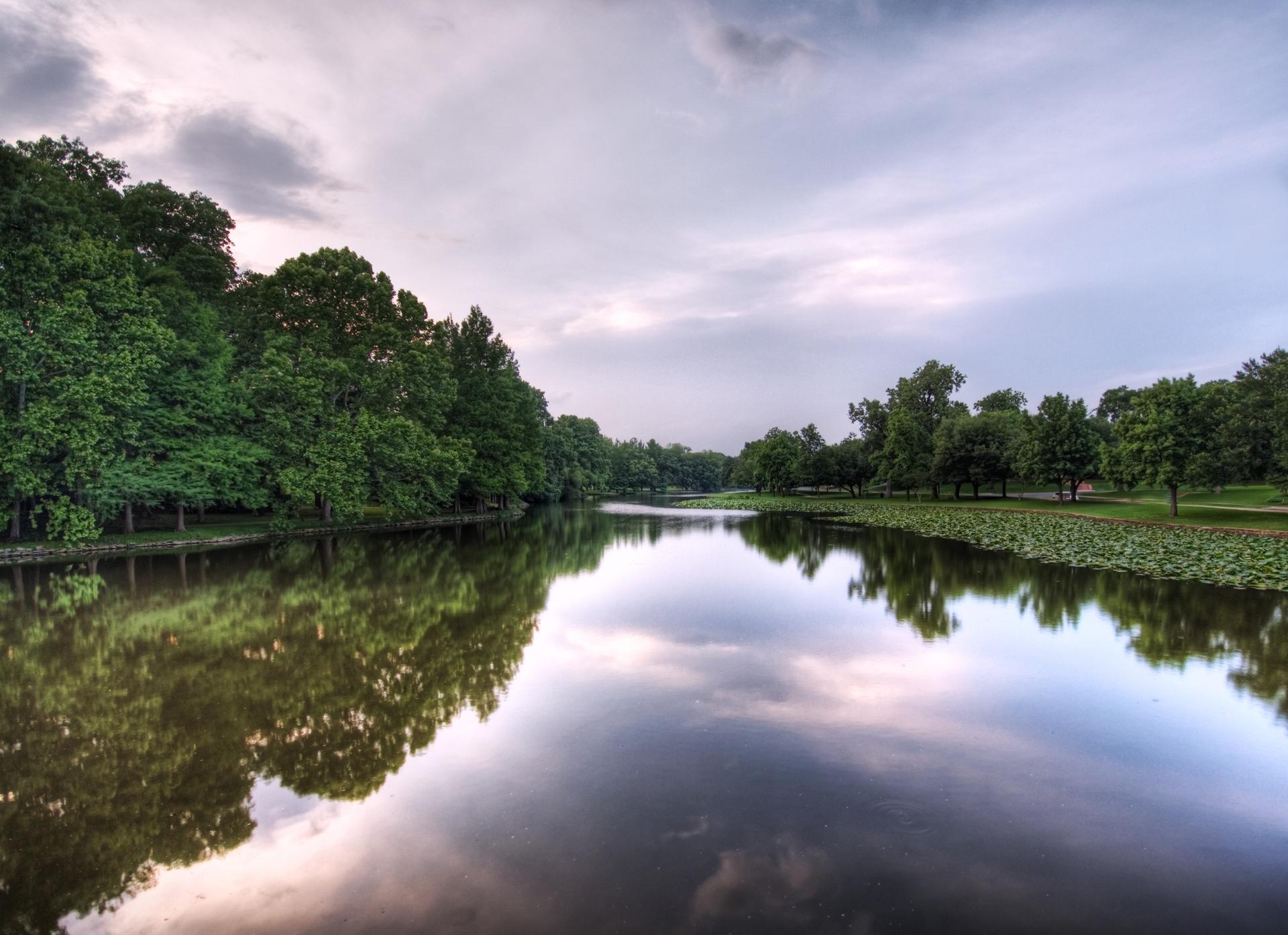 Calm river reflecting cloudy sky, flanked by lush green trees.