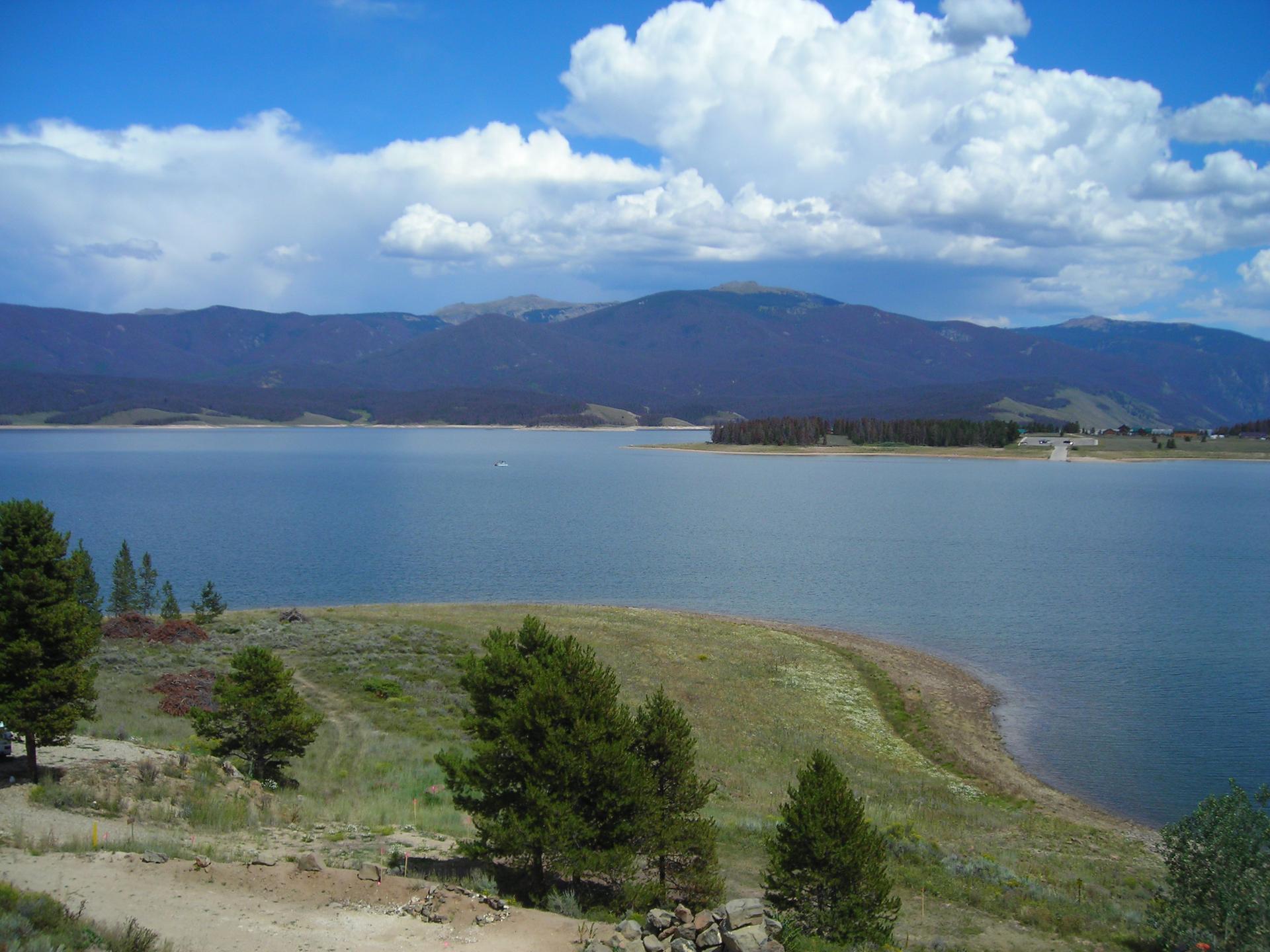 Bright daylight illuminates a large lake, distant mountains, and grassy shoreline with trees.